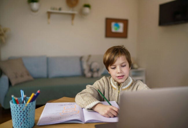 Young child doing homework in a notebook while using a laptop