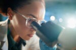 A person wearing glasses and gloves closely examining a microscope, possibly conducting scientific research in a laboratory setting.