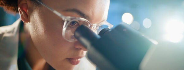 A person wearing glasses and gloves closely examining a microscope, possibly conducting scientific research in a laboratory setting.
