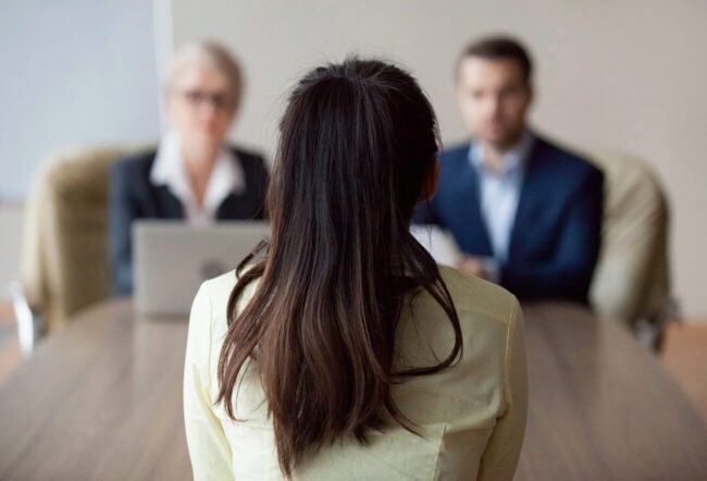 Female job candidate sitting with her back to the camera as she faces two interviewers. How do we increase the number of women in leadership?