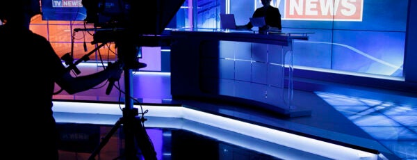 Tv news anchor sitting in his place with a camera man silhouetted in front of him.