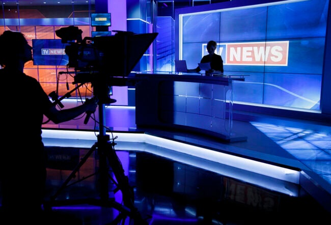 Tv news anchor sitting in his place with a camera man silhouetted in front of him.
