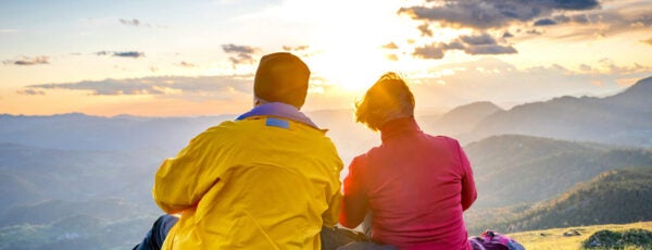 Back view of two retirees in vibrant sportswear sitting above valley and enjoying panoramic view against sunset.