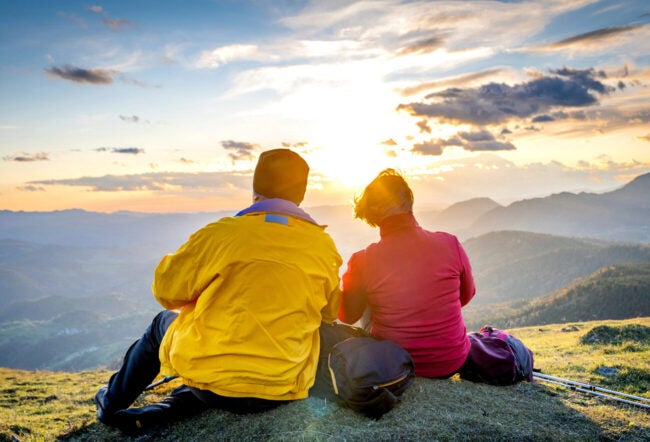 Back view of two retirees in vibrant sportswear sitting above valley and enjoying panoramic view against sunset.