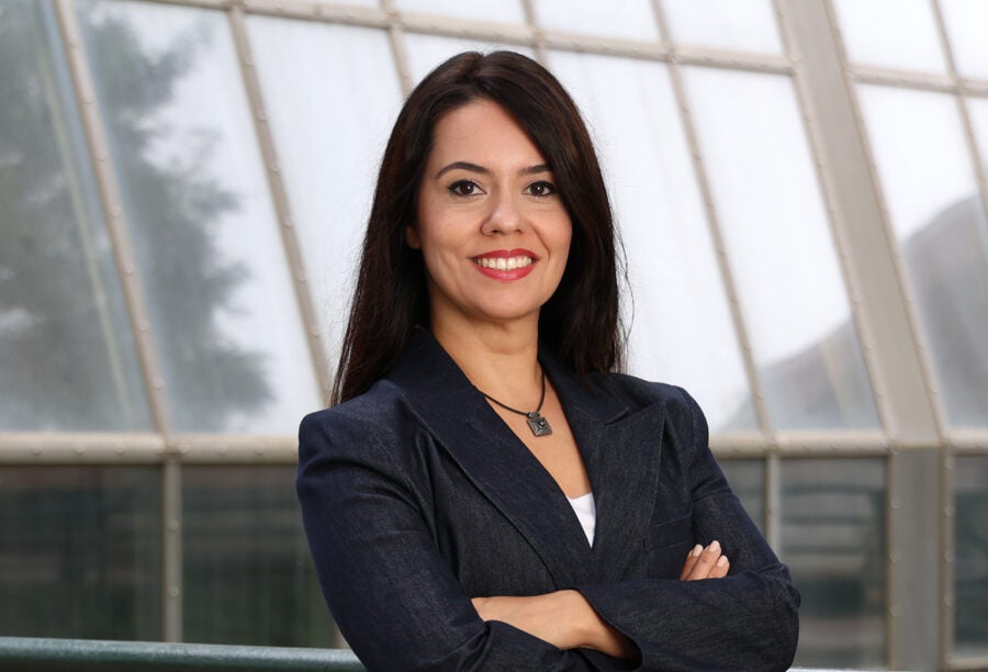 Smiling headshot of a person with long dark hair, wearing a dark blazer and necklace, standing in front of a glass-paneled building.