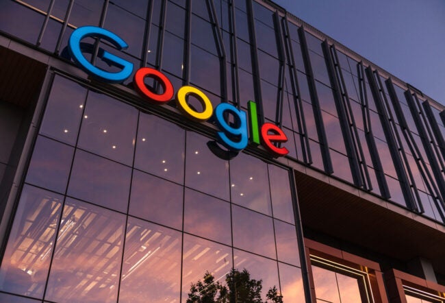 The image shows a modern glass building with the Google logo prominently displayed on the facade. The sky reflects in the windows, creating a warm ambiance.