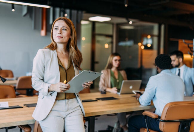 Business woman smiling and standing separate from a group of employees working in the background