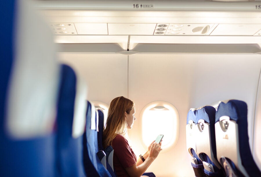 A person is sitting in an airplane seat by the window, using a smartphone. The interior of the plane is visible, with rows of blue seats and overhead compartments. Sunlight is coming through the window.