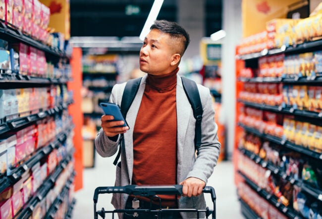 A person with a backpack pushes a shopping cart down a supermarket aisle, holding a smartphone and looking at the shelves.