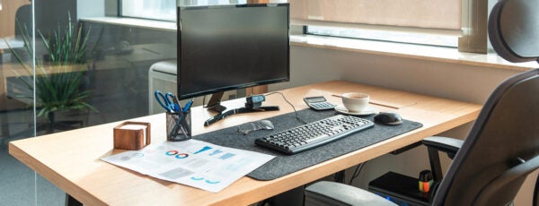 An organized office desk with a computer monitor, keyboard, mouse, and a cup of coffee. Documents, a calculator, and stationery are also present. A window provides natural light.