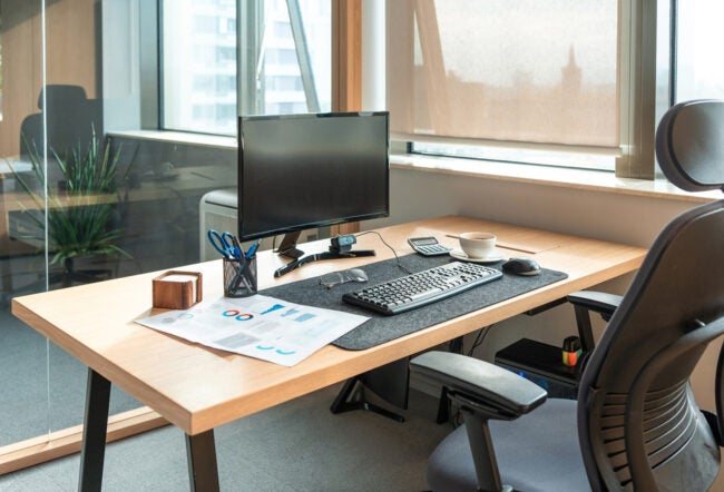 An organized office desk with a computer monitor, keyboard, mouse, and a cup of coffee. Documents, a calculator, and stationery are also present. A window provides natural light.