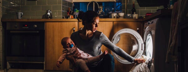 Young mother is holding her crying baby daughter on her hip while trying to load the washing machine.