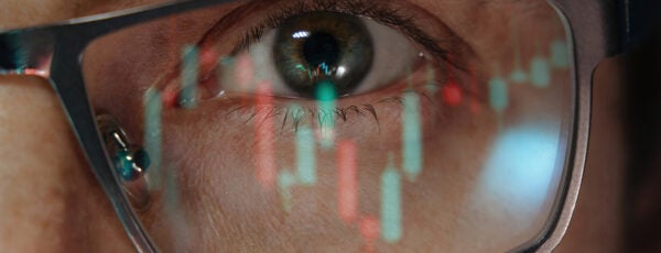A close-up of an eye wearing glasses with a stock market chart reflection, representing financial analysis or market focus.
