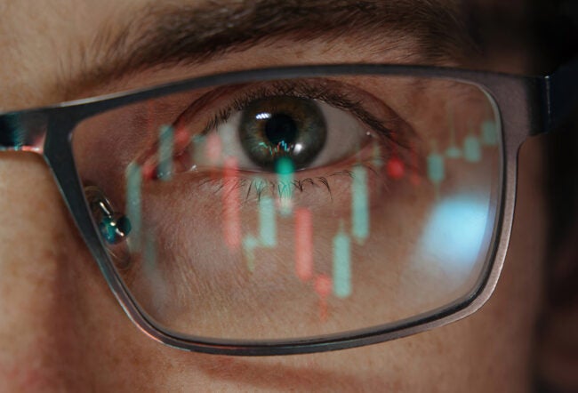 A close-up of an eye wearing glasses with a stock market chart reflection, representing financial analysis or market focus.
