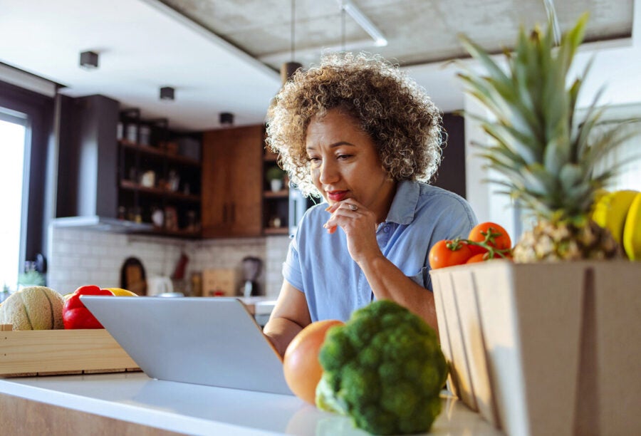 Woman using her laptop in the kitchen while surrounded by fresh groceries and produce in the kitchen