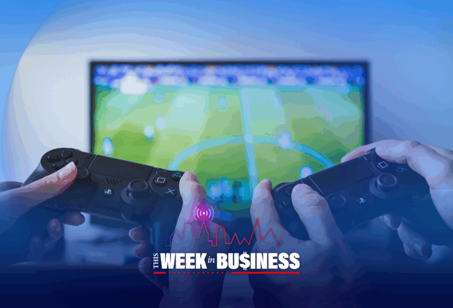 A few people holding video game controllers in front of a TV screen displaying a soccer game. Overlay text reads "This Week in Business" with a city skyline and graph.