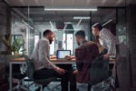 Three people in a modern office environment collaborating around a laptop on a conference table.