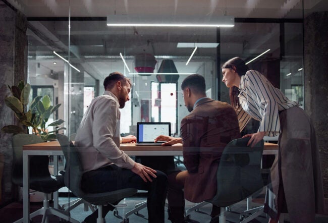 Three people in a modern office environment collaborating around a laptop on a conference table.