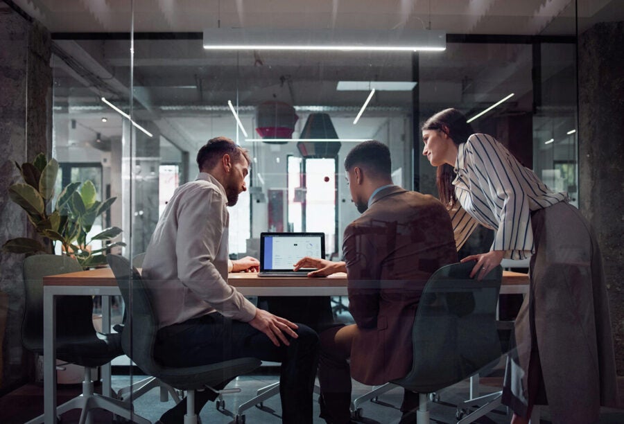 Three people in a modern office environment collaborating around a laptop on a conference table.