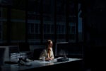 A person working late at night in a dimly lit office, illuminated by a computer monitor.