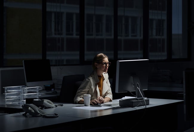 A person working late at night in a dimly lit office, illuminated by a computer monitor.