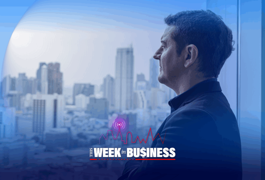 Person looking out a window at a cityscape, with the text "This Week in Business" overlaying the foreground. The scene conveys a professional, contemplative mood.