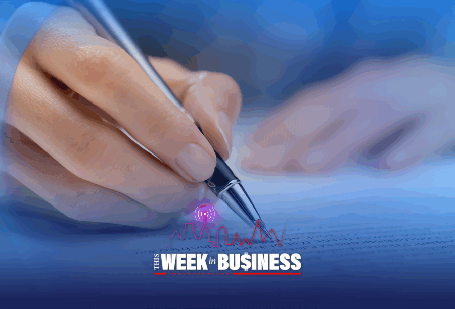 A hand holding a pen, writing on a page filled with text. In the foreground, the text "The Week in Business" is displayed with a chart line and signal icon, suggesting a business or financial theme.