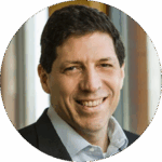 Headshot of person smiling, wearing a suit jacket and collared shirt, with blurred background.